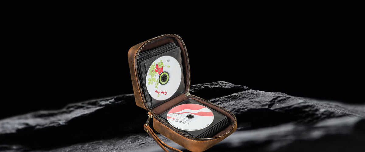 A brown leather CD case shown open, containing two CDs, set against a dark, textured rock surface with a solid black background.