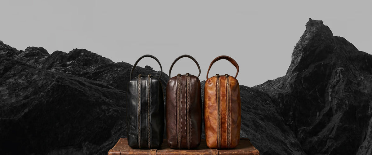 Three leather travel or duffel bags in black, dark brown, and tan are lined up on a wooden crate against a dramatic, dark mountain backdrop.