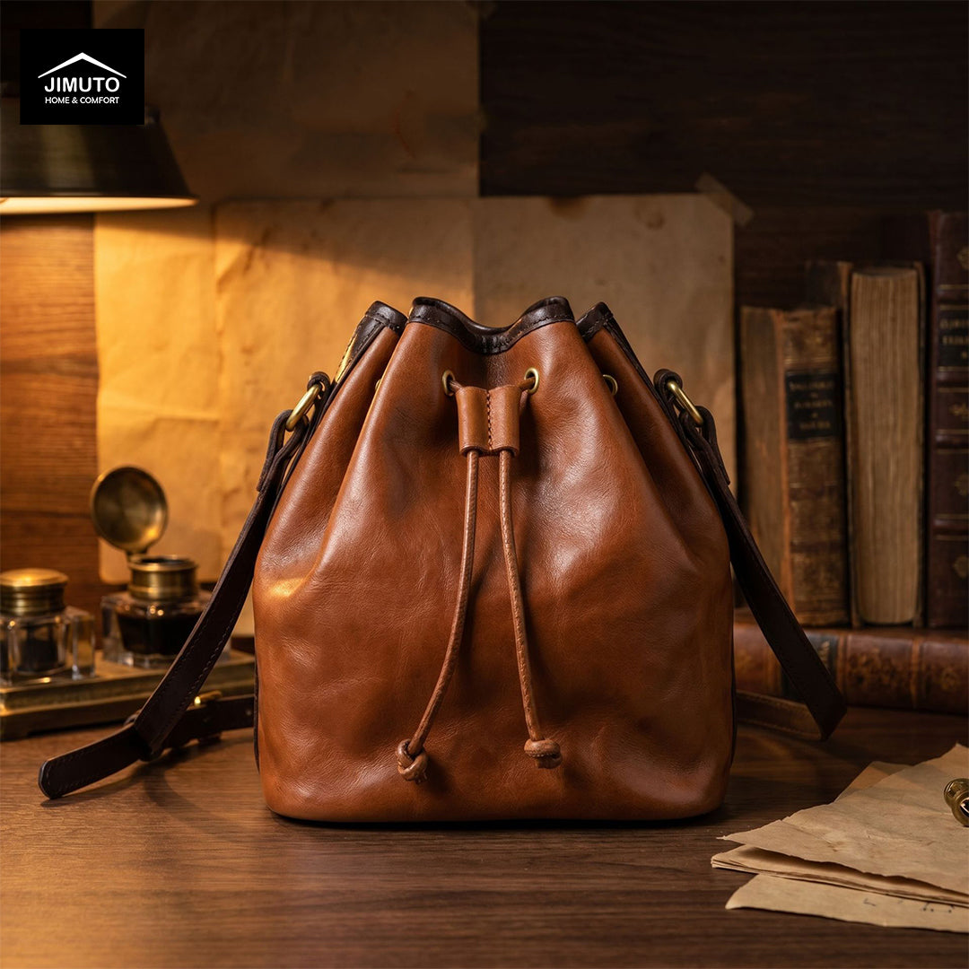 A brown top-grain leather bucket bag with a drawstring closure and black shoulder strap, displayed on a wooden table in a warm, vintage-styled room with books and a lamp. The Jimuto brand logo is visible in the top-left corner.