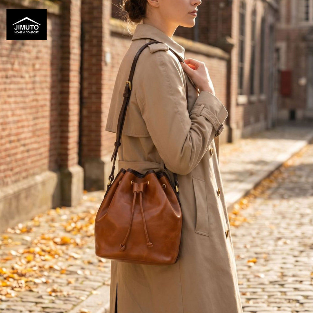 A woman in a beige trench coat carries a brown TO-GRAIN leather bucket bag with a black strap and gold-tone hardware, photographed outdoors on a cobblestone street with brick buildings in the background. The Jimuto brand logo appears in the top-left corner.
