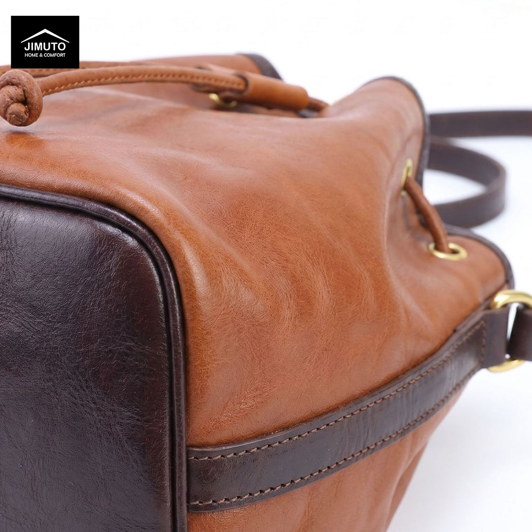 A close-up of a JIMUTO leather bucket bag showing a warm tan front panel, a dark brown side panel, a dark brown strap, and gold-tone hardware on a white background.