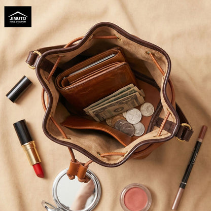 An open brown top-grain leather bucket bag filled with a wallet, cash, and coins, styled on a beige fabric background with cosmetics around it. The Jimuto brand logo appears in the top-left corner.