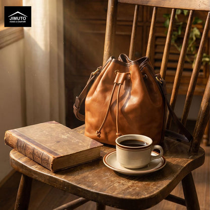 A tan leather bucket bag with a drawstring closure sits on a rustic wooden chair, styled with an old book and a cup of coffee. The Jimuto Designs logo is visible in the top-left corner.