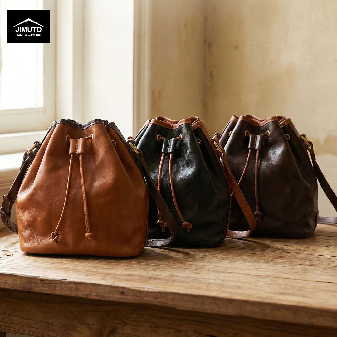 Three leather bucket bags in tan, black, and dark brown are lined up on a rustic wooden table in a warm, sunlit room. The JIMUTO brand logo is visible in the top-left corner.