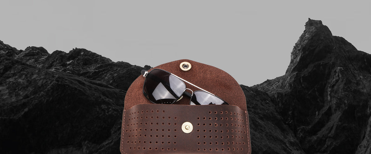 A brown leather sunglasses case with gold-tone hardware, shown open with a pair of sunglasses inside, set against a dark mountain landscape and gray sky.