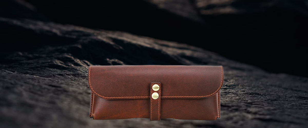 A brown leather eyeglasses case with gold-tone snap buttons, displayed on a dark, textured rock surface.