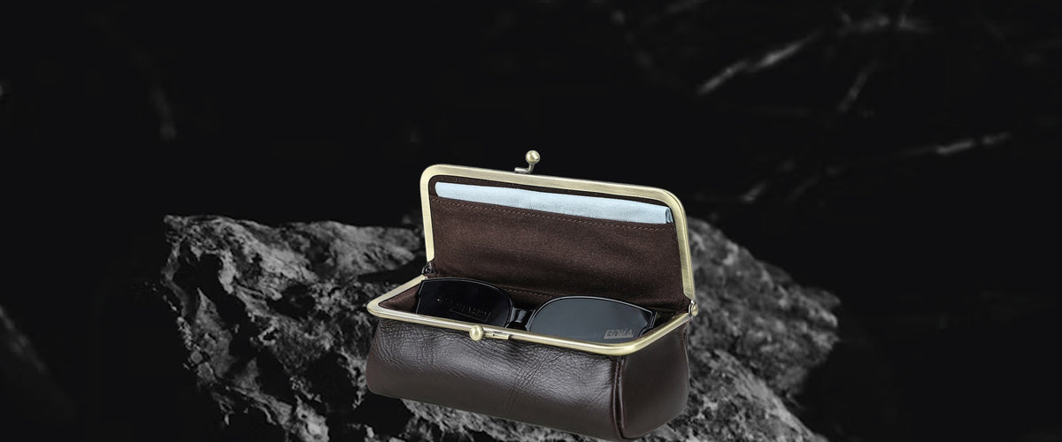 A dark brown leather sunglasses case with gold-tone metal hardware, shown open on a dark textured rock surface against a black background.