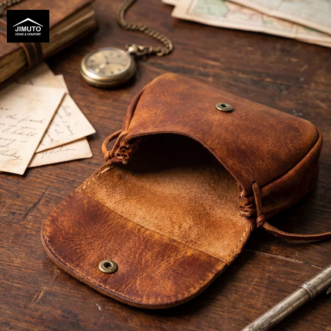 A brown leather pouch or small bag with an open flap, photographed on a rustic wooden tabletop with vintage props including a pocket watch, papers, and a pen. A Jimuto logo appears in the top-left corner.