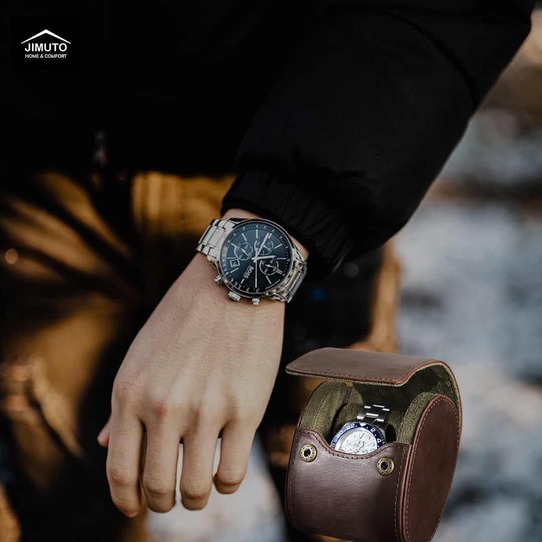 A silver-toned stainless steel watch with a black dial is worn on a person's wrist, with a brown leather watch pouch holding another watch nearby. The background is softly blurred, and the Jimuto logo appears in the top-left corner.