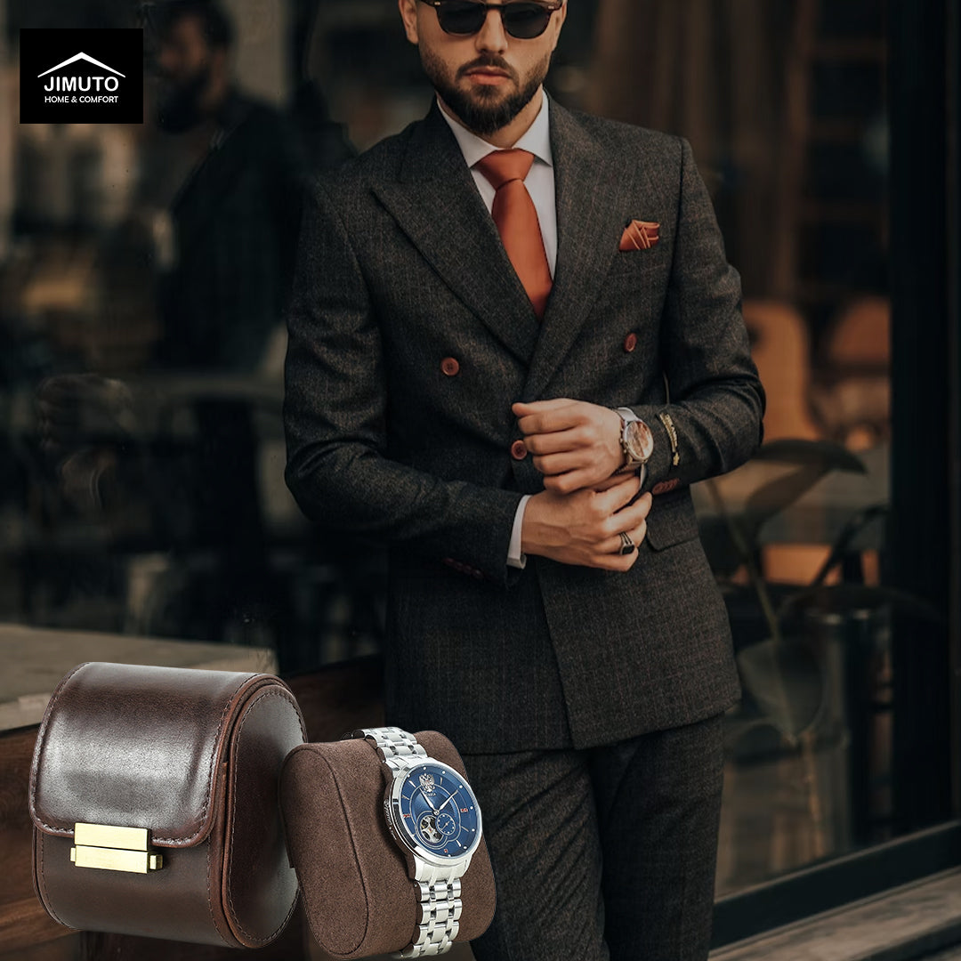 A man wearing a dark gray double-breasted suit, orange tie, and sunglasses stands in front of a window. A brown leather watch box and a wristwatch with a blue dial are displayed in the foreground.