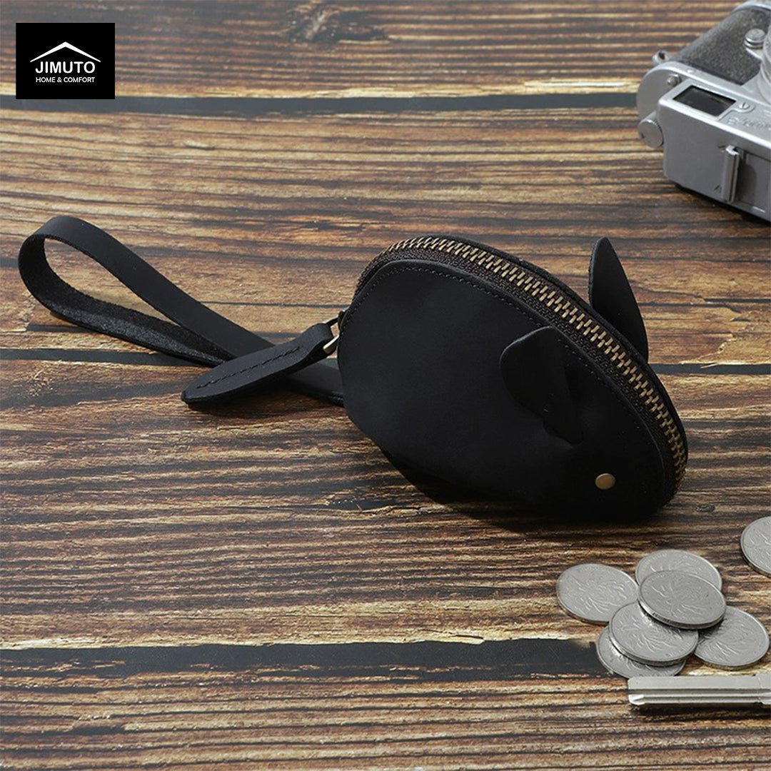 A black top-grain leather mouse-shaped zippered coin purse with a wrist strap, displayed on a rustic wooden surface alongside coins, a key, and a vintage camera. The Jimuto brand logo is visible in the top-left corner.