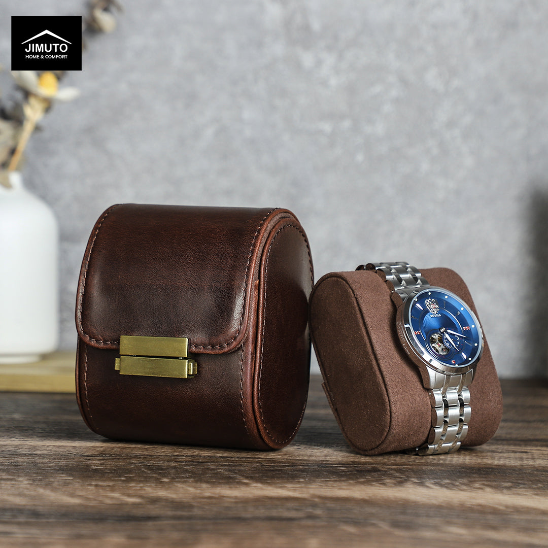 A dark brown leather watch box with a gold clasp and a matching brown suede-like watch cushion, shown with a wristwatch on a wooden surface against a gray wall. The Jimuto brand logo is visible in the top left corner.