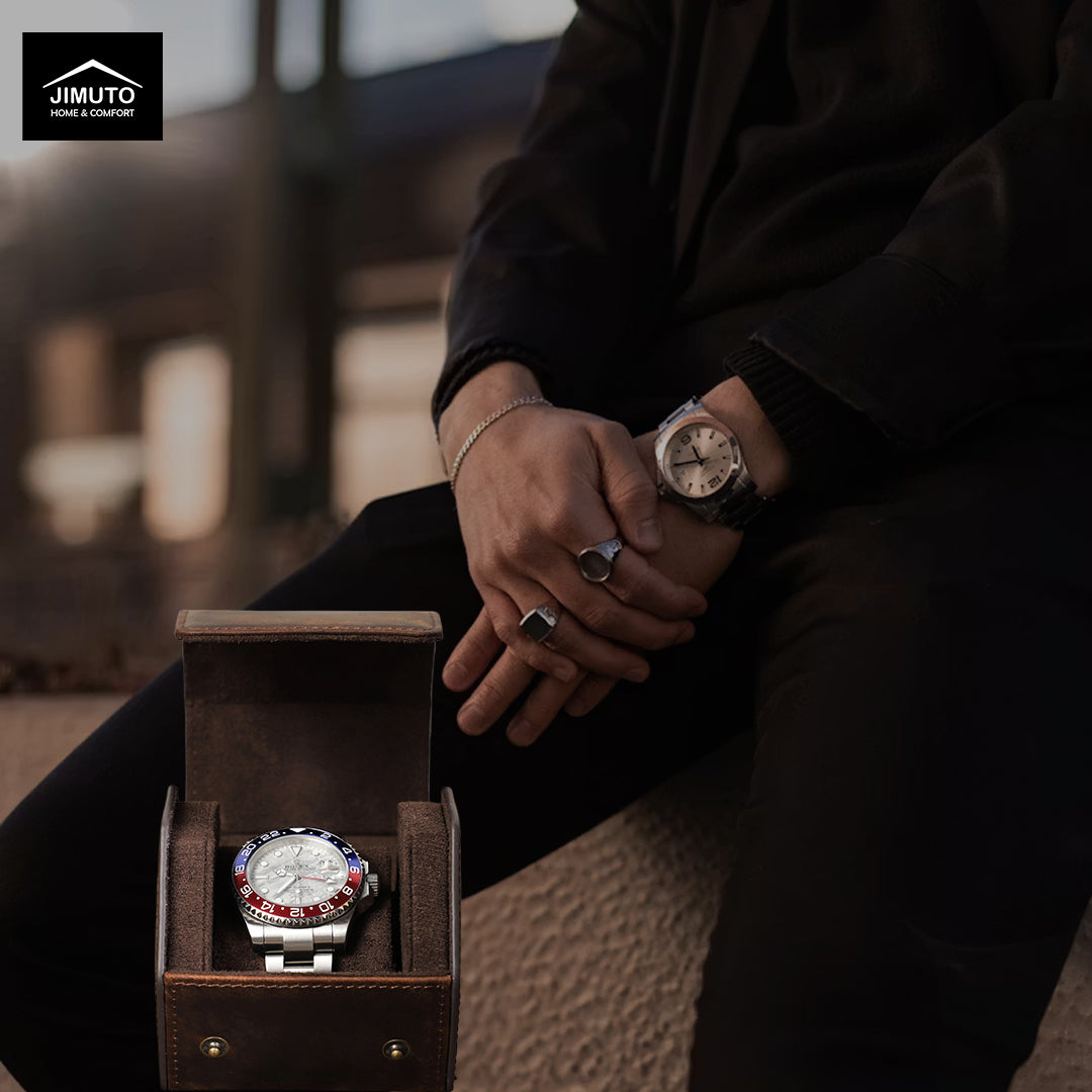 A person in a black suit sits with hands crossed, wearing a wristwatch, while an open brown hexagonal leather watch box holding another matching watch rests beside them. The scene is softly lit with a blurred outdoor background, and the Jimuto logo appears in the top-left corner.