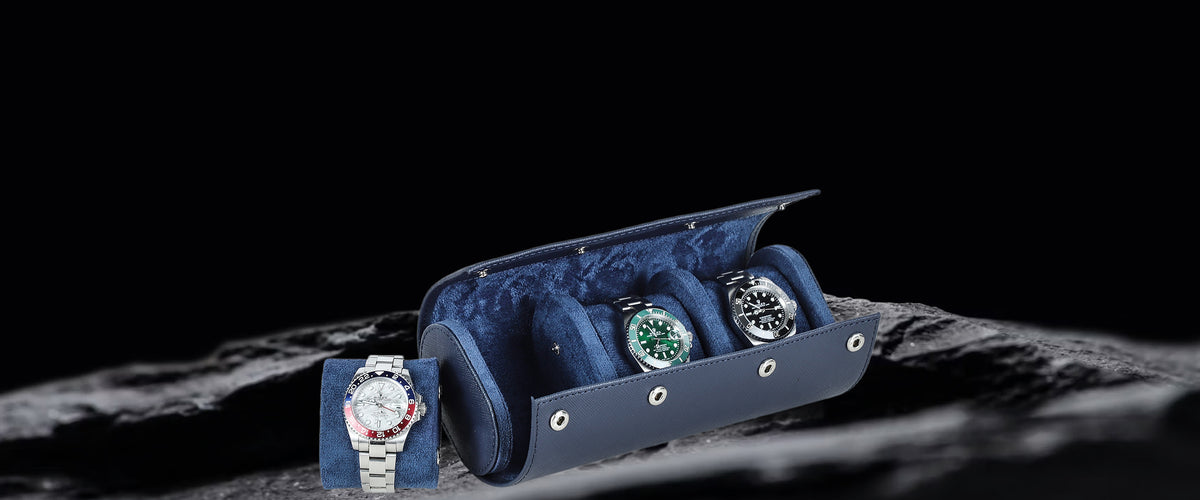 A navy blue Saffiano Leather Watch Storage Box with three watch slots, shown open on a dark textured surface. A wristwatch with a silver-toned case and white dial is displayed outside the case on the left, and two additional watches are visible inside the slots.