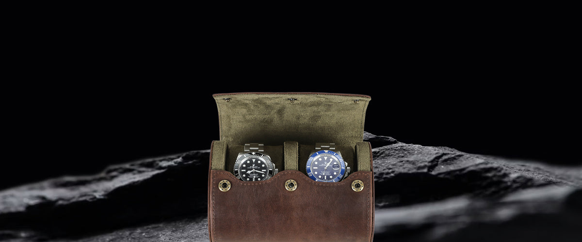 A brown crazy horse leather watch box with a hinged lid, shown open on a dark textured surface against a black background. Two watches are partially visible inside: one with a black dial and bezel, and one with a blue dial and bezel.