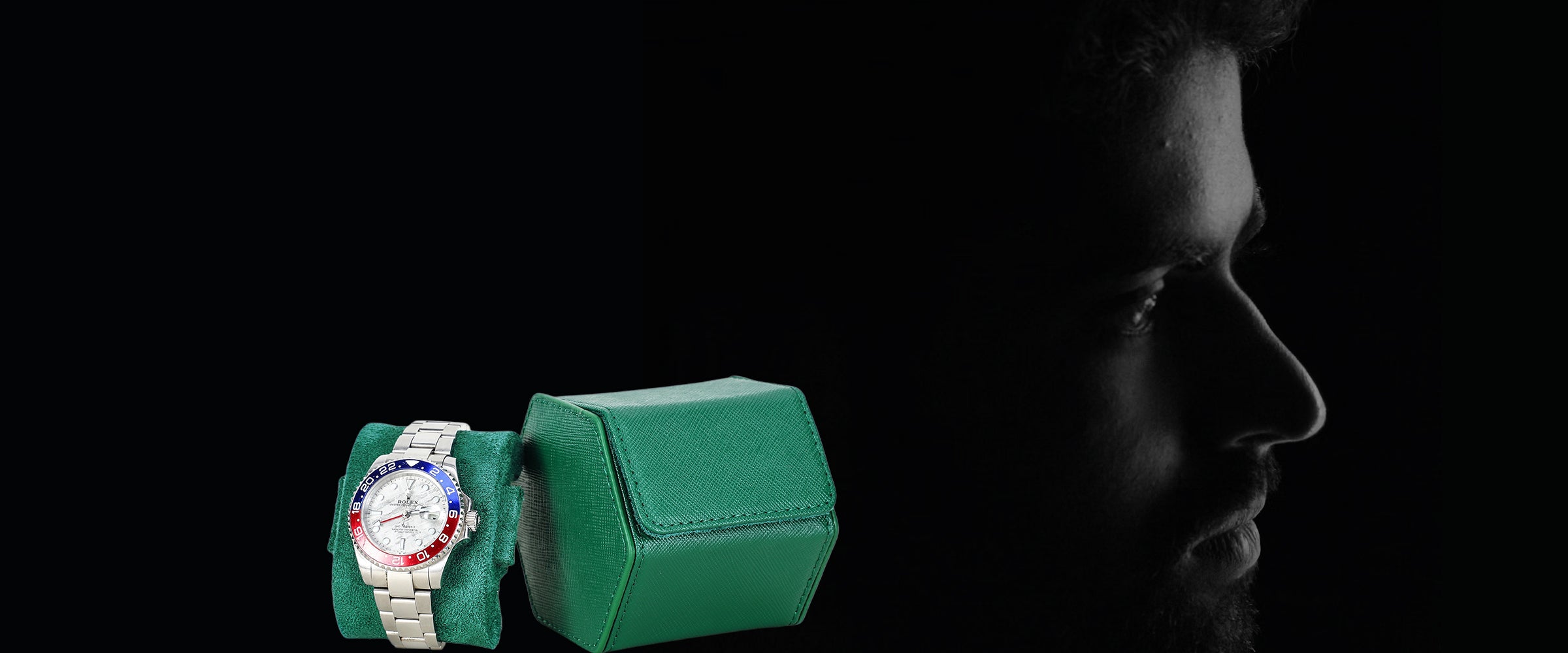A wristwatch with a silver-toned metal bracelet and a red, white, and black bezel is displayed on a green cushion next to a green saffiano leather watch box, set against a dark background with a silhouetted man's profile on the right.