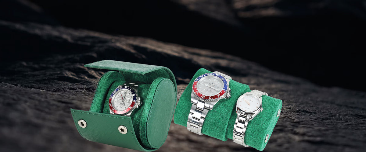 Two silver metal wristwatches displayed on green watch cushions, with one watch inside an open green Saffiano Leather Watch Box and the other on a matching cushion, set against a dark textured background.
