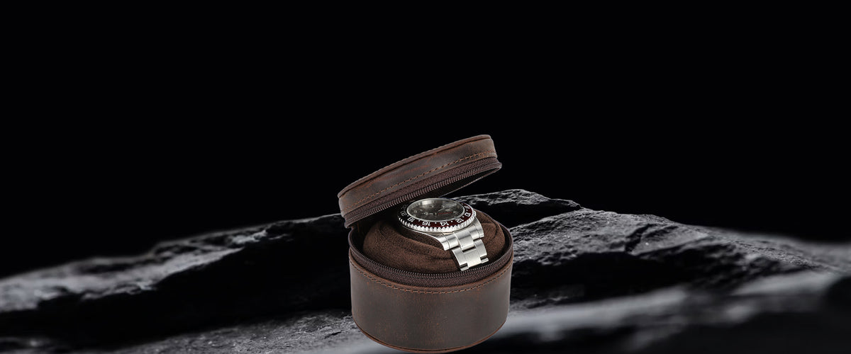 A silver-toned stainless steel watch with a dark dial sits inside an open cylindrical brown crazy horse leather watch box, placed on a dark textured rock against a black background.