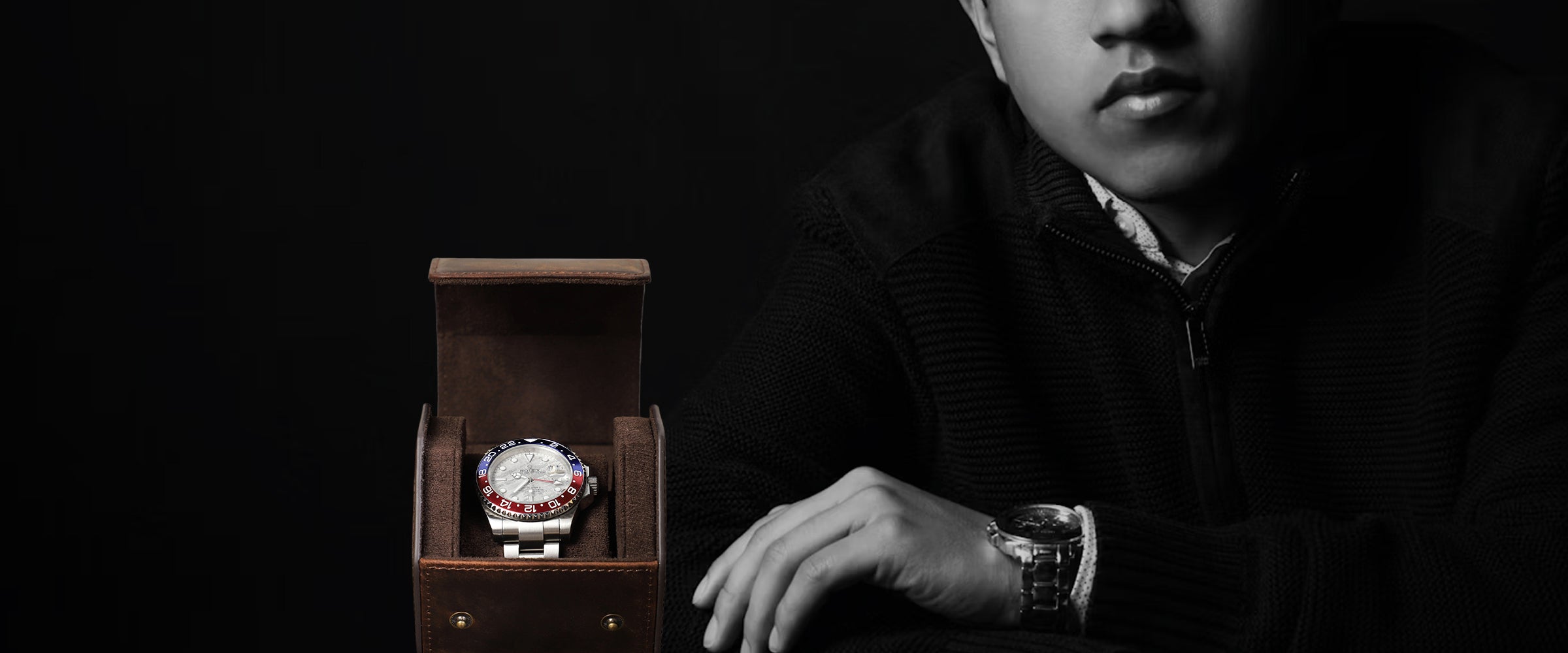 A wristwatch with a white dial and red bezel detail displayed inside an open brown leather watch box, with a person in a dark sweater partially visible on the right against a black background.