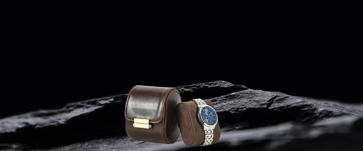 A luxury watch with a blue dial and silver-toned bracelet displayed next to its brown leather watch case on a dark textured rock surface against a black background.
