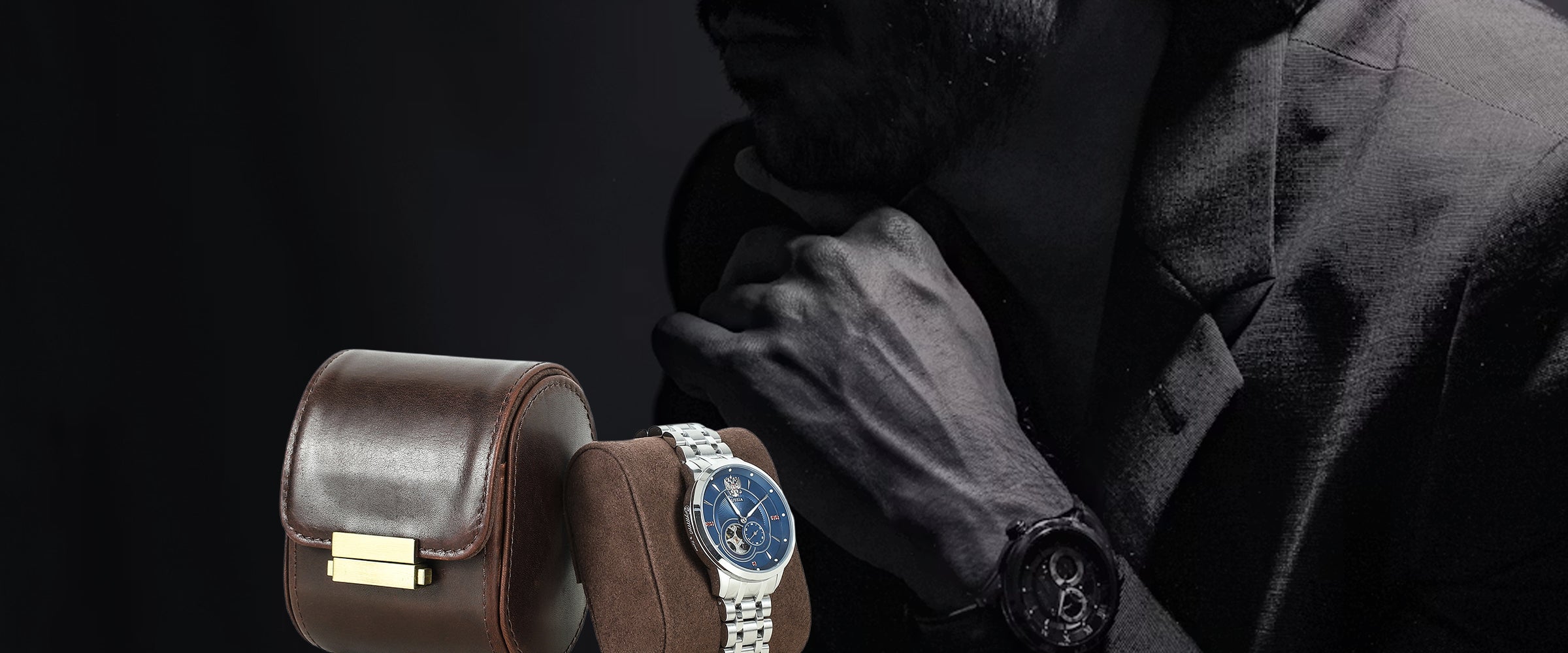 A luxury watch with a silver-toned metal bracelet and a deep blue dial is displayed on a brown leather watch cushion, with a man in a dark suit wearing a black watch in the background against a dark studio backdrop.