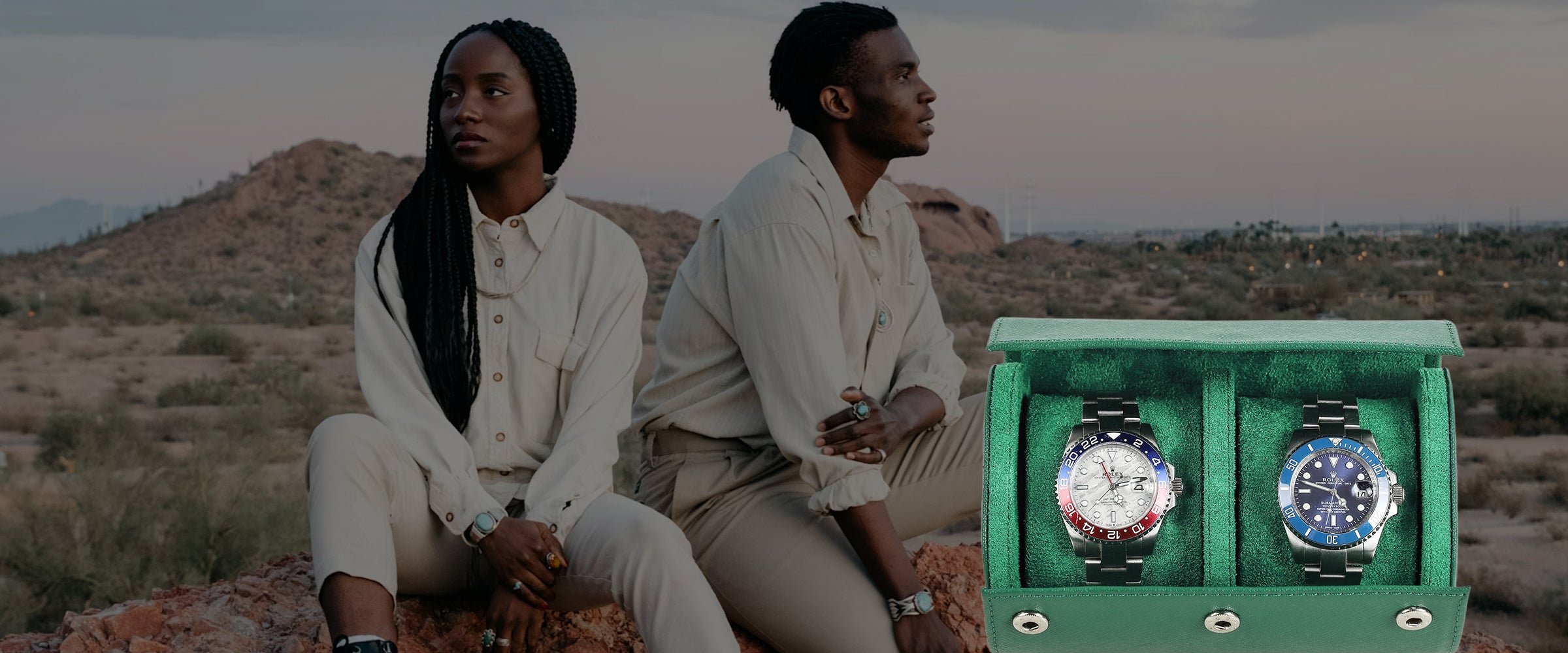 Two people sitting in a desert landscape with a green saffiano watch case containing two watches.