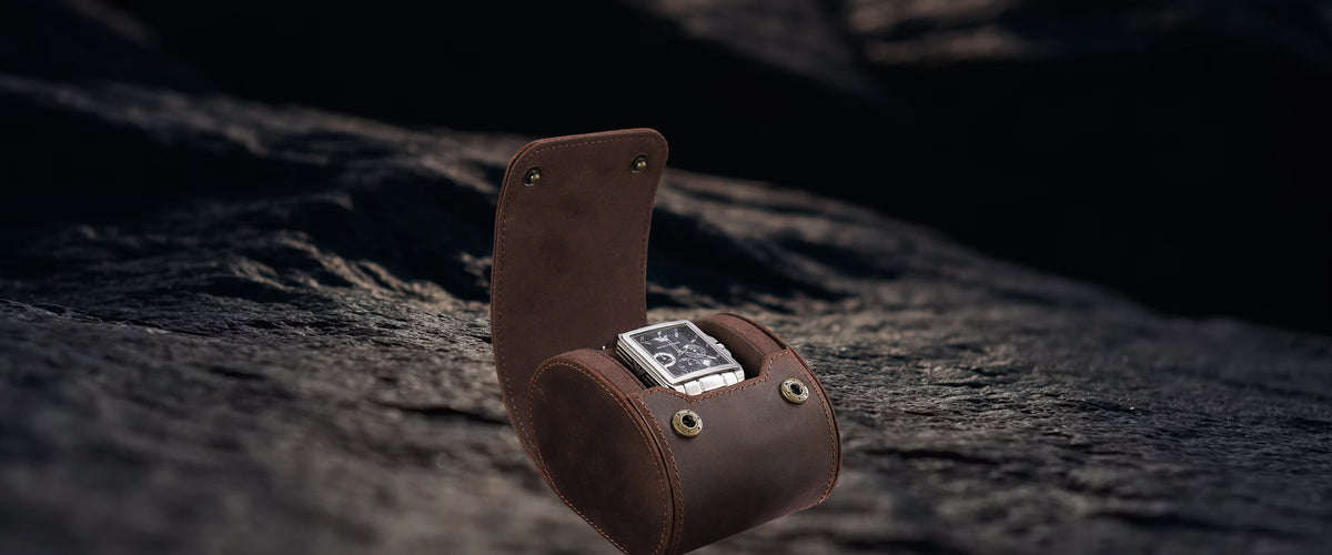 A brown crazy horse leather watch pouch holding a silver-toned watch, set against a dark textured rock background.