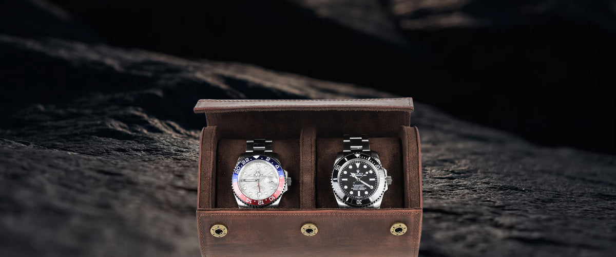 Two luxury wristwatches displayed side-by-side inside an open brown crazy horse leather watch box on a dark, textured stone surface.