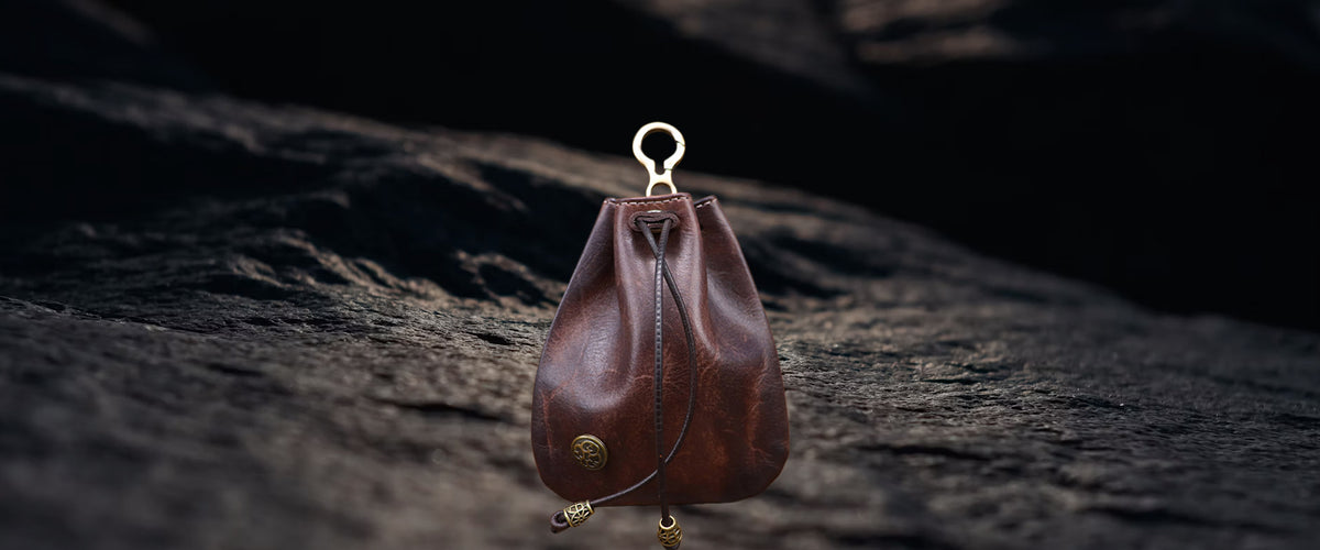 A small brown leather drawstring pouch bag with brass hardware, hanging against a dark textured rock background.