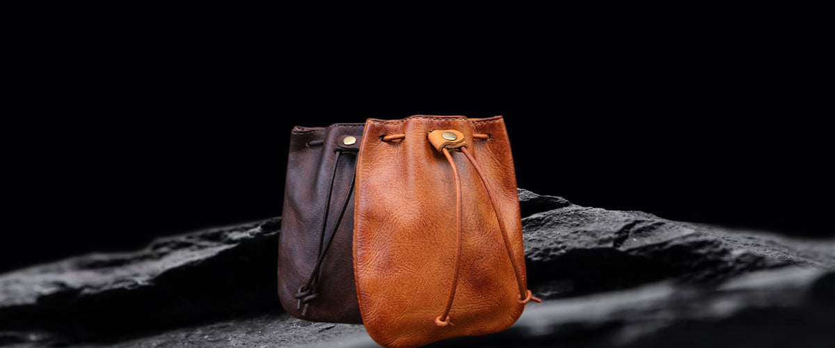 Two leather drawstring pouches or small bags standing upright on a dark textured surface against a black background. The front bag is a warm cognac brown, and the rear bag is a darker brown.