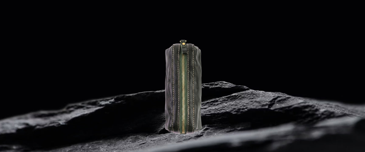 A slim, structured black leather pen pouch with vertical gold-tone chain detailing, standing upright on a dark textured surface against a solid black background.