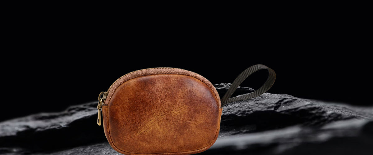 A small oval brown leather pouch with a zipper closure and wrist strap, displayed on a dark textured surface against a black background.
